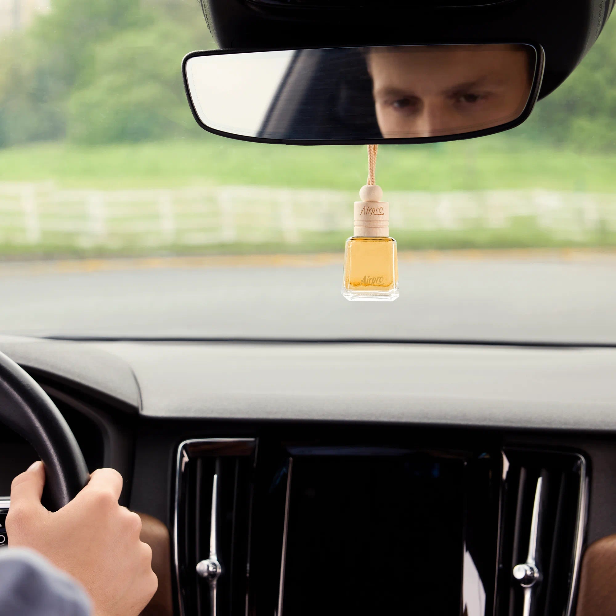 An elegant Airpro Bloom Dew glass bottle car diffuser with a wooden cap hanging gracefully from a car rearview mirror during a drive.