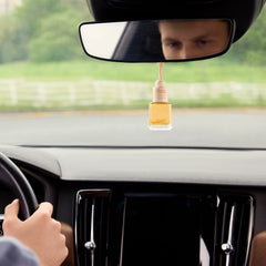 An elegant Airpro Bloom Dew glass bottle car diffuser with a wooden cap hanging gracefully from a car rearview mirror during a drive.
