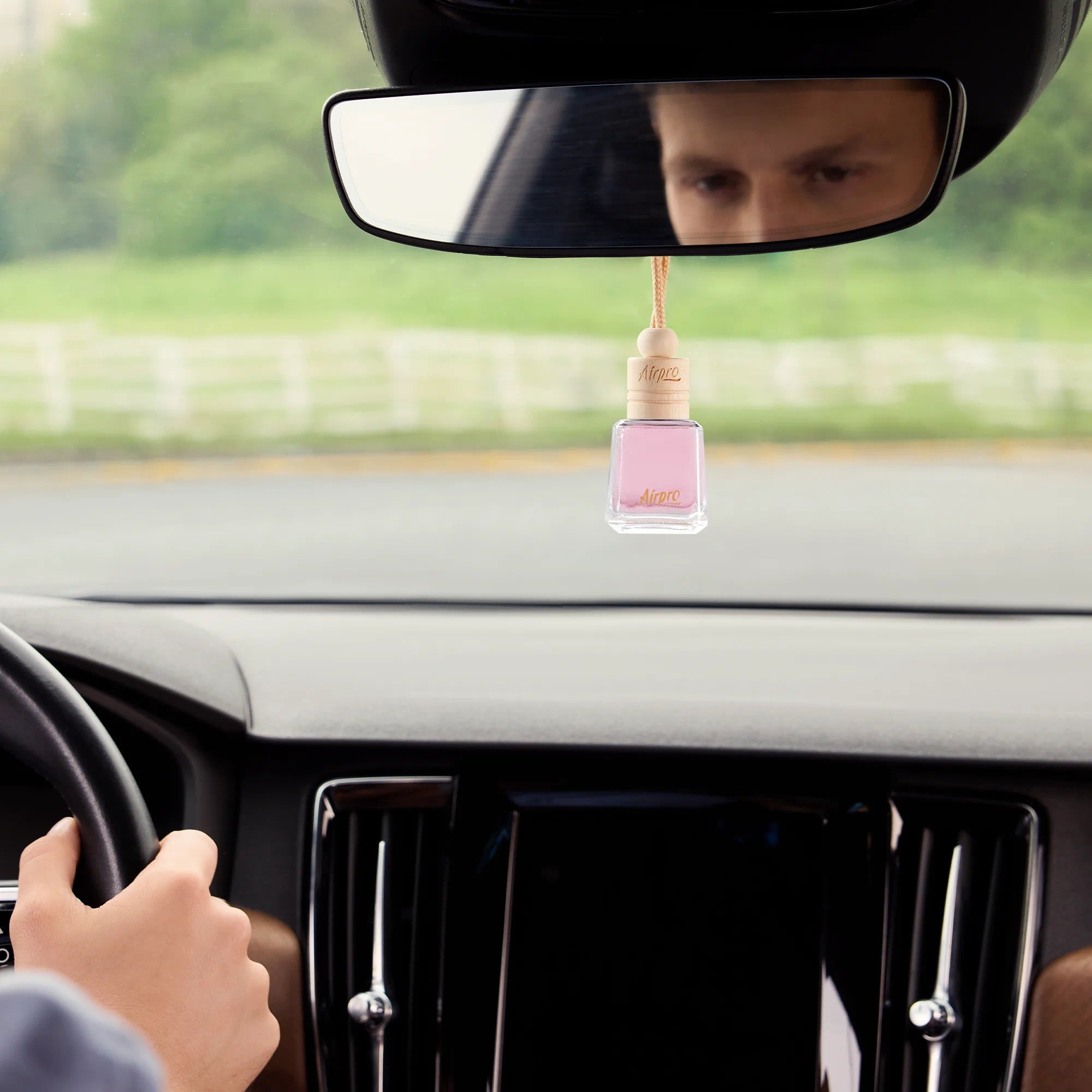 An Airpro floral Sakura Lumi glass bottle car scent with a wooden cap hanging from a car rearview mirror during a drive.