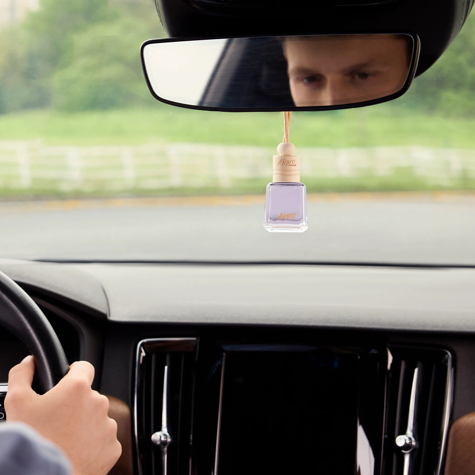 An elegant Airpro Velvet Mist glass bottle car freshener with a wooden cap hanging from a car rearview mirror during a daylight drive.